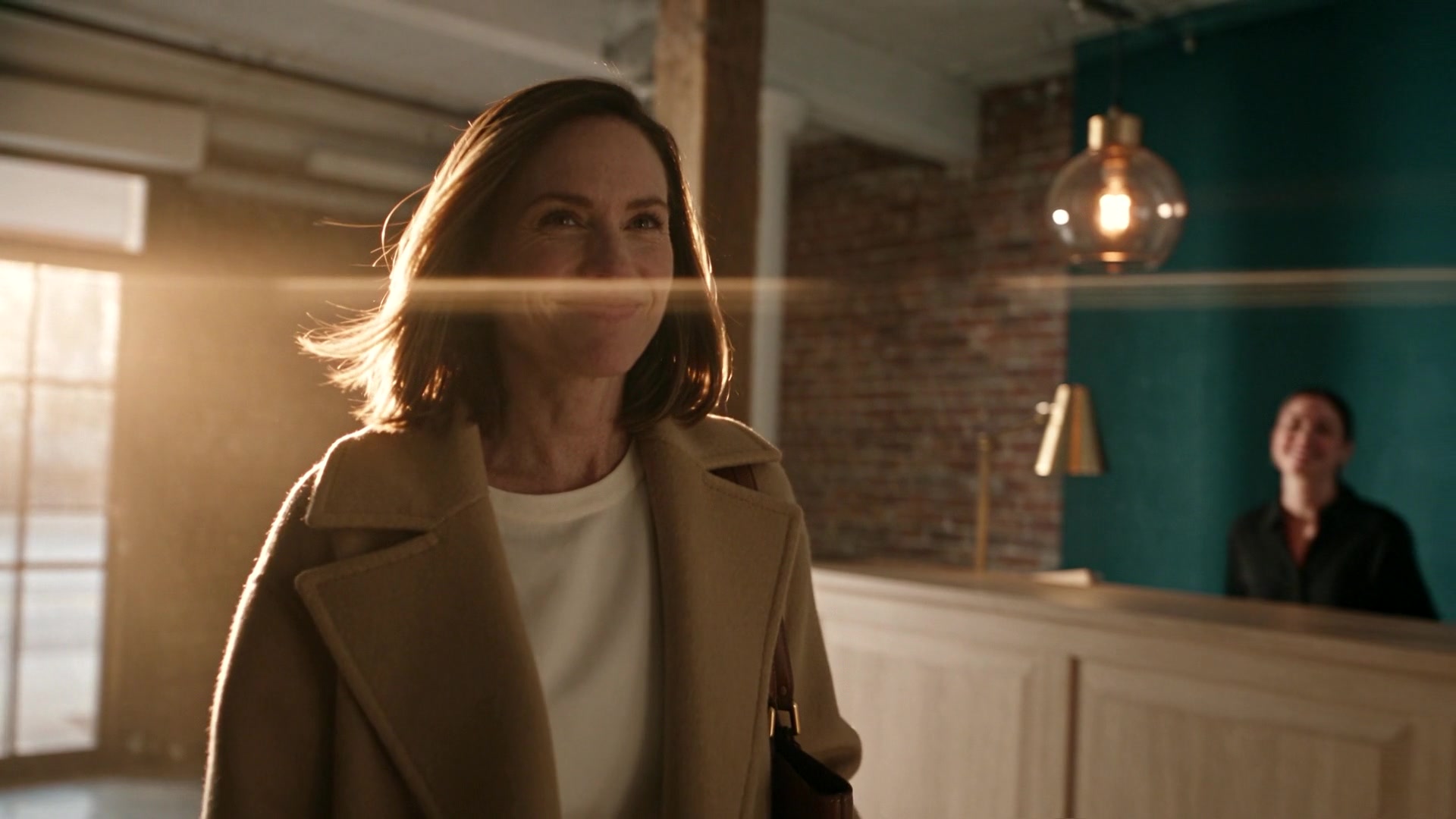 A smiling woman in a camel coat arriving at a reception desk, a receptionist behind it