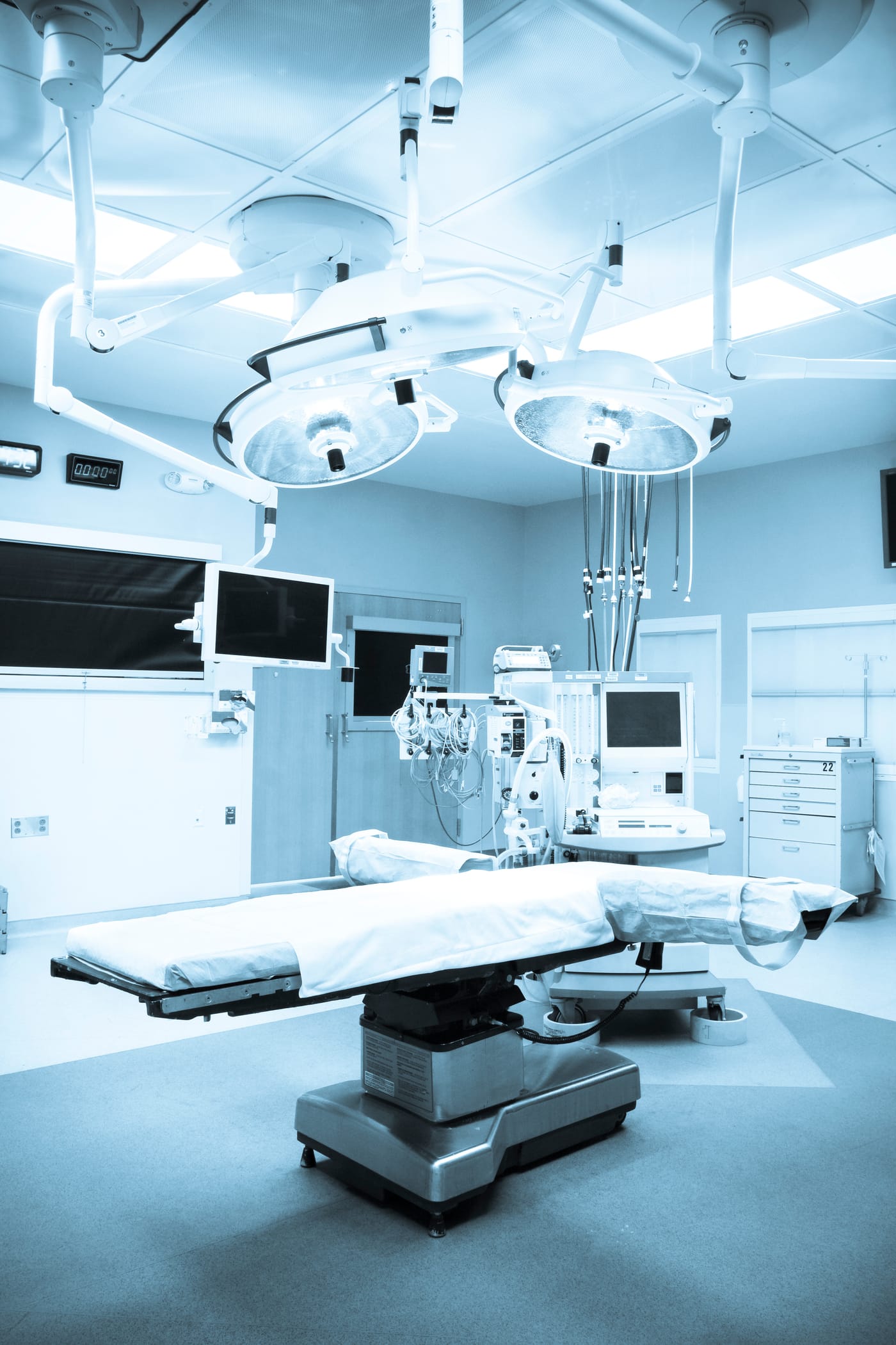 An empty operating room under surgical lights, the table made up and the monitors on