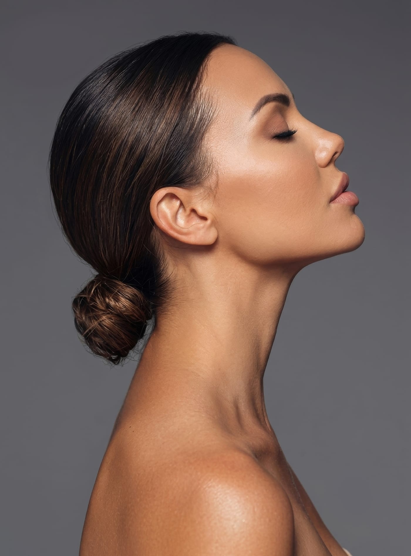 A model in profile with her dark hair in a low bun, eyes closed, against a grey background