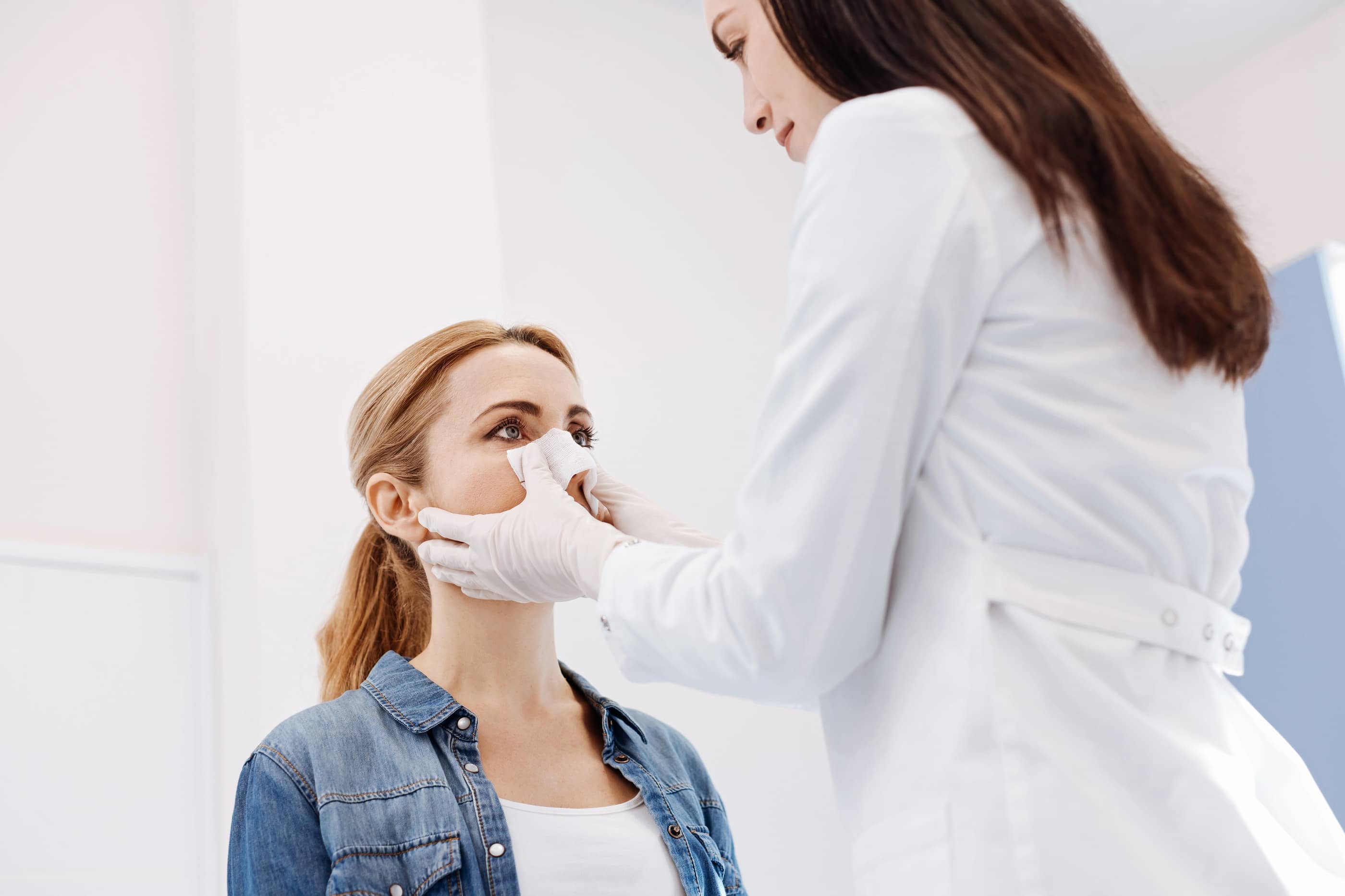 A doctor in a white coat gently touching the nose of a young woman in a denim shirt
