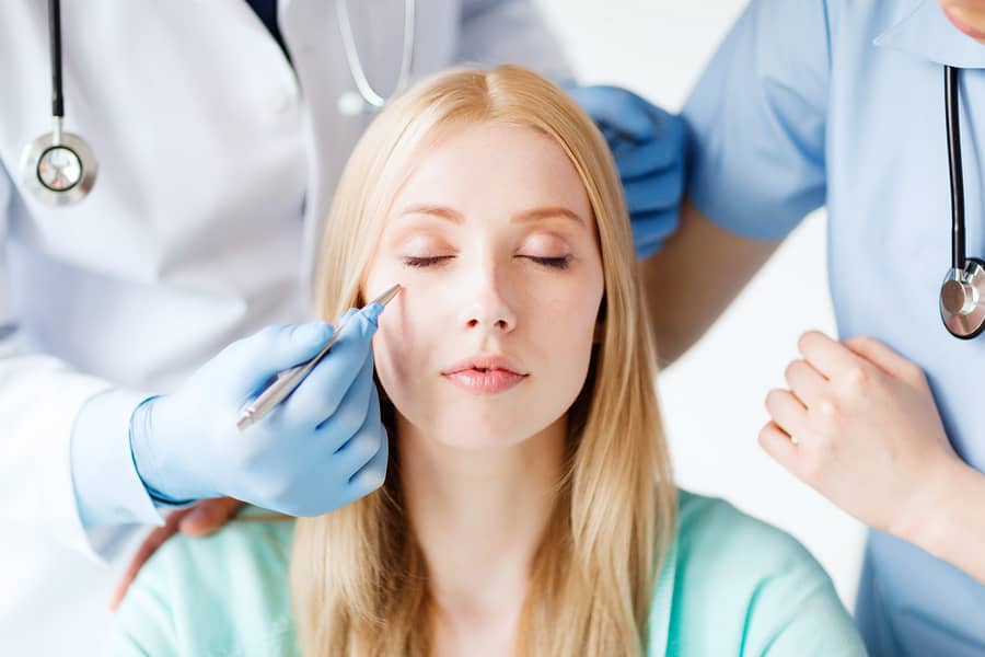 A woman with closed eyes being marked on the face with a pen by a gloved doctor, a second clinician beside them